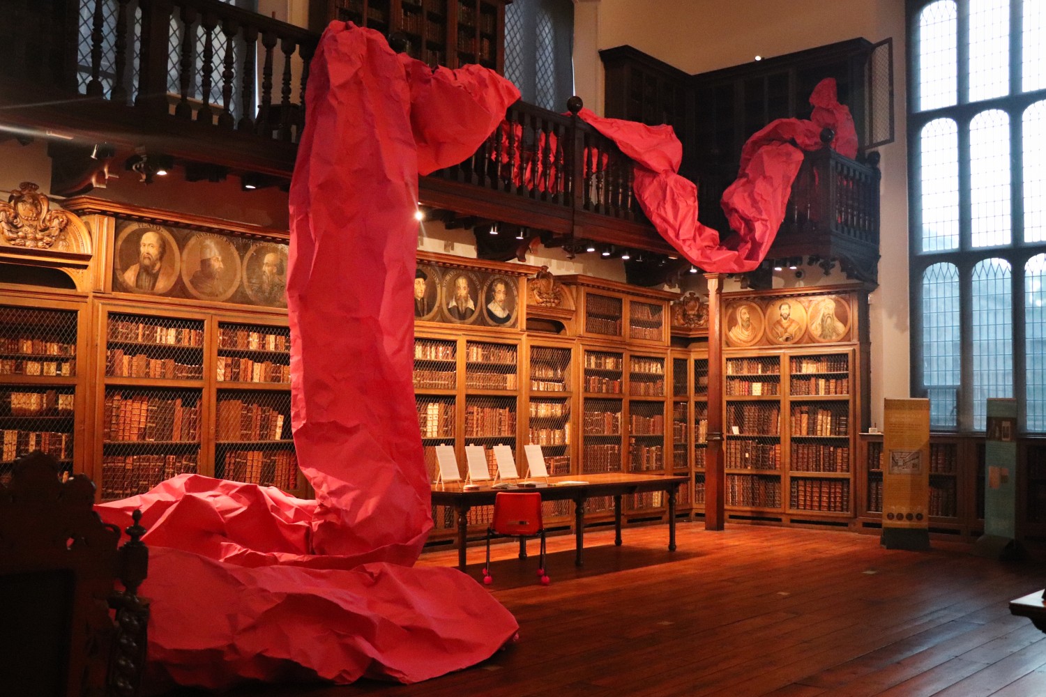 image description: colour photo, interior. 17th century building. a library with a wooden balcony running around the inside of the room around 10 feet from the floor. there are bookshelves on all walls below and above the balcony containing 1000s of beautiful old books. across around two thirds of the balcony and down onto the floor oozes a vast ribbon of bright pink paper. on the floor envelops by the swirling end of the paper is a larger bright orange bean bag where viewer can sit and experience the installation from the inside.