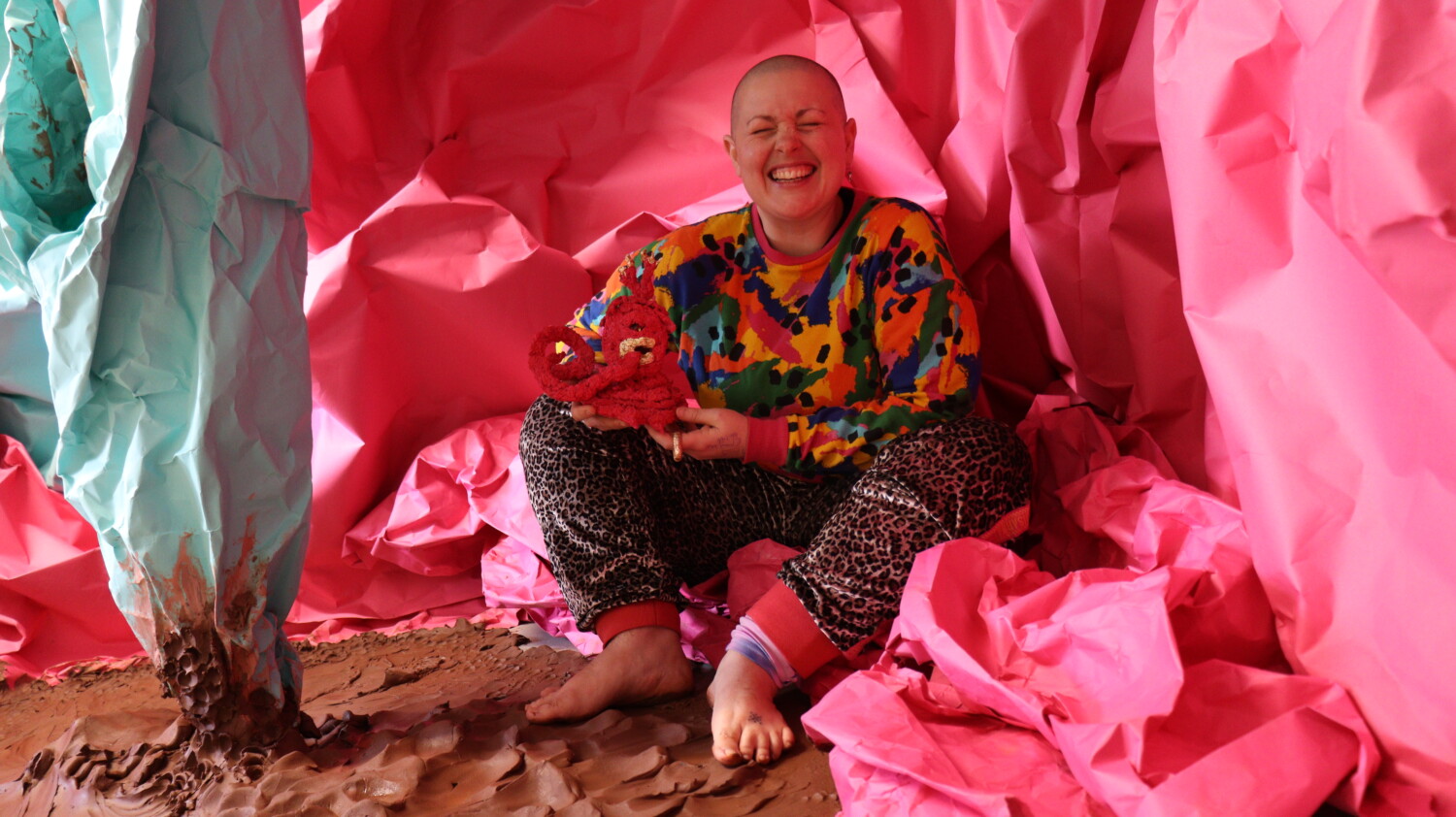 Colour photo, interior. Kitt, a white, shaven headed person with a wide crinkly nosed smile, sits amongst vast sheets of pink and mint green paper crumple to form a cave - like structure around them. on the floor is wet, raw terracotta clay in rolling folds. Credit: Art Matters Now
