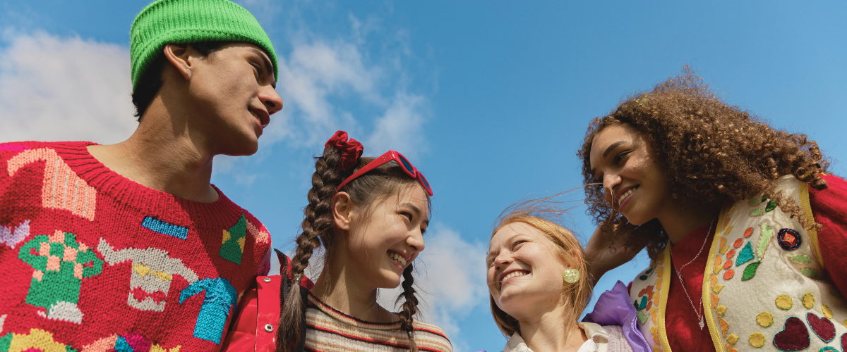Colour image taken from below of the faces of young people talking to each other looking happy. The young people, of various ages, are engaged with each other. The sky is blue.